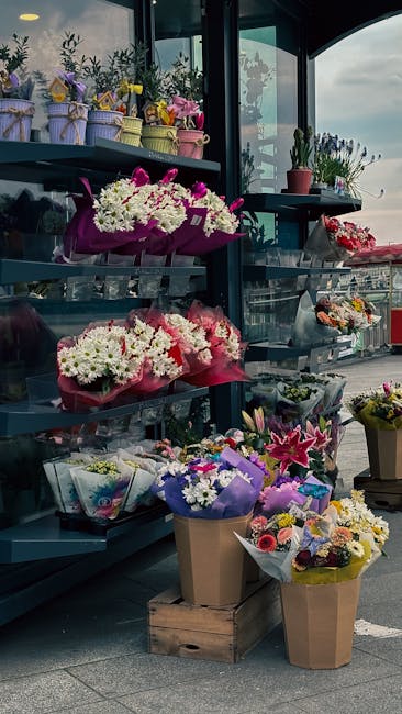 A display of vibrant floral arrangements on metal shelves inside a shop with large glass windows revealing an outdoor view. The flowers include white daisies, pink lilies, and purple blossoms, arrange