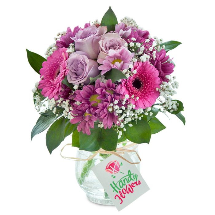Taking pride of place in this vivid image is a lush, rounded bouquet featuring a lively mix of pinks, purples, and hints of fresh green, perfectly capturing the creative spirit of Poplar. At its heart, soft lavender roses with elegantly curling petals set a gentle, romantic tone, reminiscent of fragrant gardens tucked away behind Poplar's Victorian terraces. These are ringed by blushing fuchsia Gerbera daisies, their dramatic, dark-centered faces beaming outward in friendly exuberance, perfectly balanced by clusters of deep magenta chrysanthemums with luminous, yellow-green eyes. Sprigs of delicate, snowy baby's breath are woven through the design, lending an airy brightness that recalls early morning light streaming through local shop windows. Lush, healthy green leaves frame the blooms and peep out from unexpected corners, their glossy surface adding nuanced texture and a sense of fresh abundance. The arrangement rests inside a clear glass vase, notable for its distinctive crackled finish that glimmers softly in the light-a hallmark of local Poplar craftsmanship. Tied around the neck is a natural raffia ribbon and a crisp white tag, inviting a handwritten message or special greeting-making this bouquet as fitting a gift for a birthday near Poplar Recreation Ground as for a housewarming along Abbott Road. This thoughtfully gathered bouquet speaks to the unique artistry and warmth of local flower delivery in Poplar.